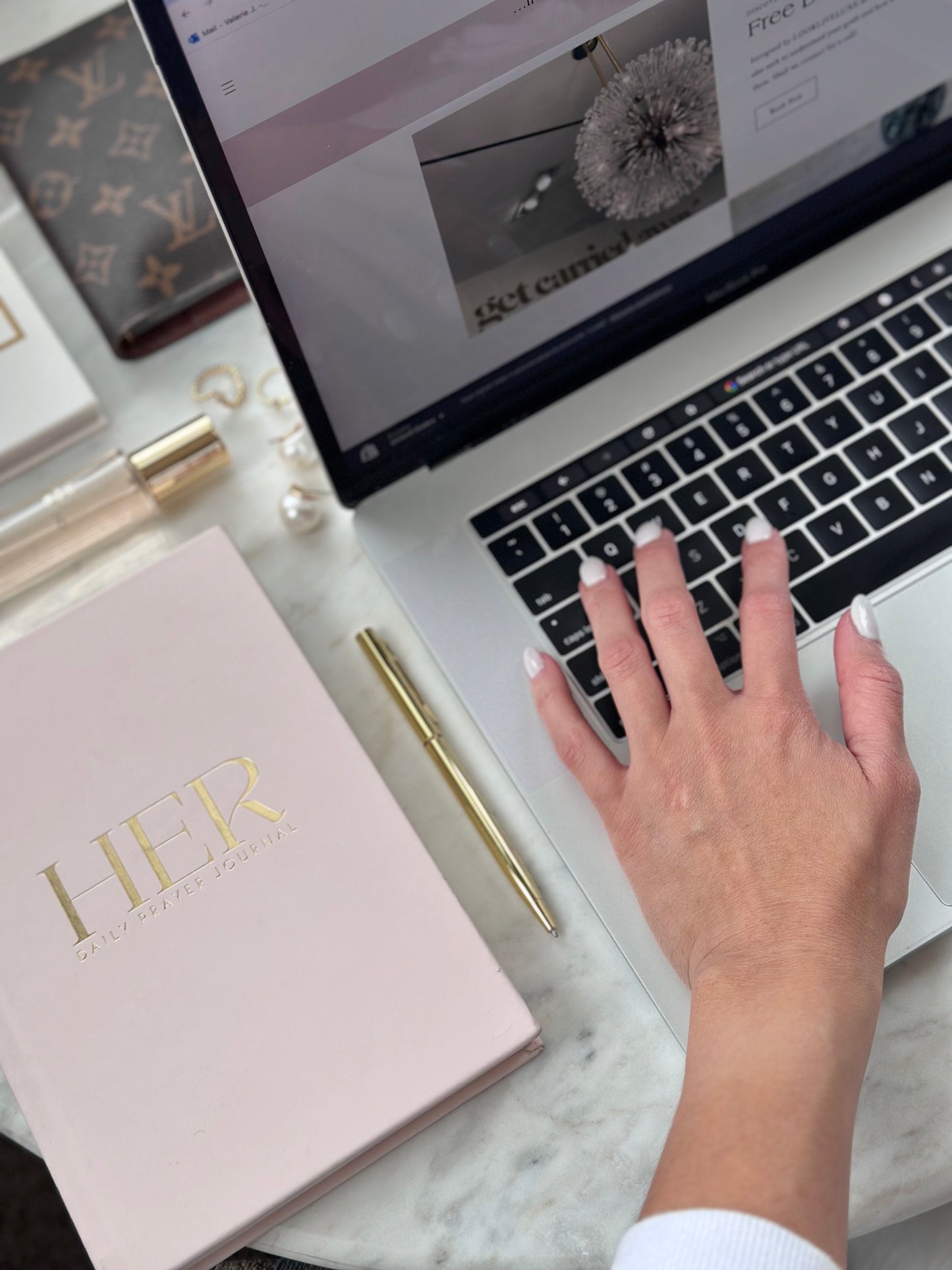 Valerie's hand using a laptop with a luxury brand's product in the foreground, including a pink notebook with gold lettering and a gold pen at LOOKLIVELUXE